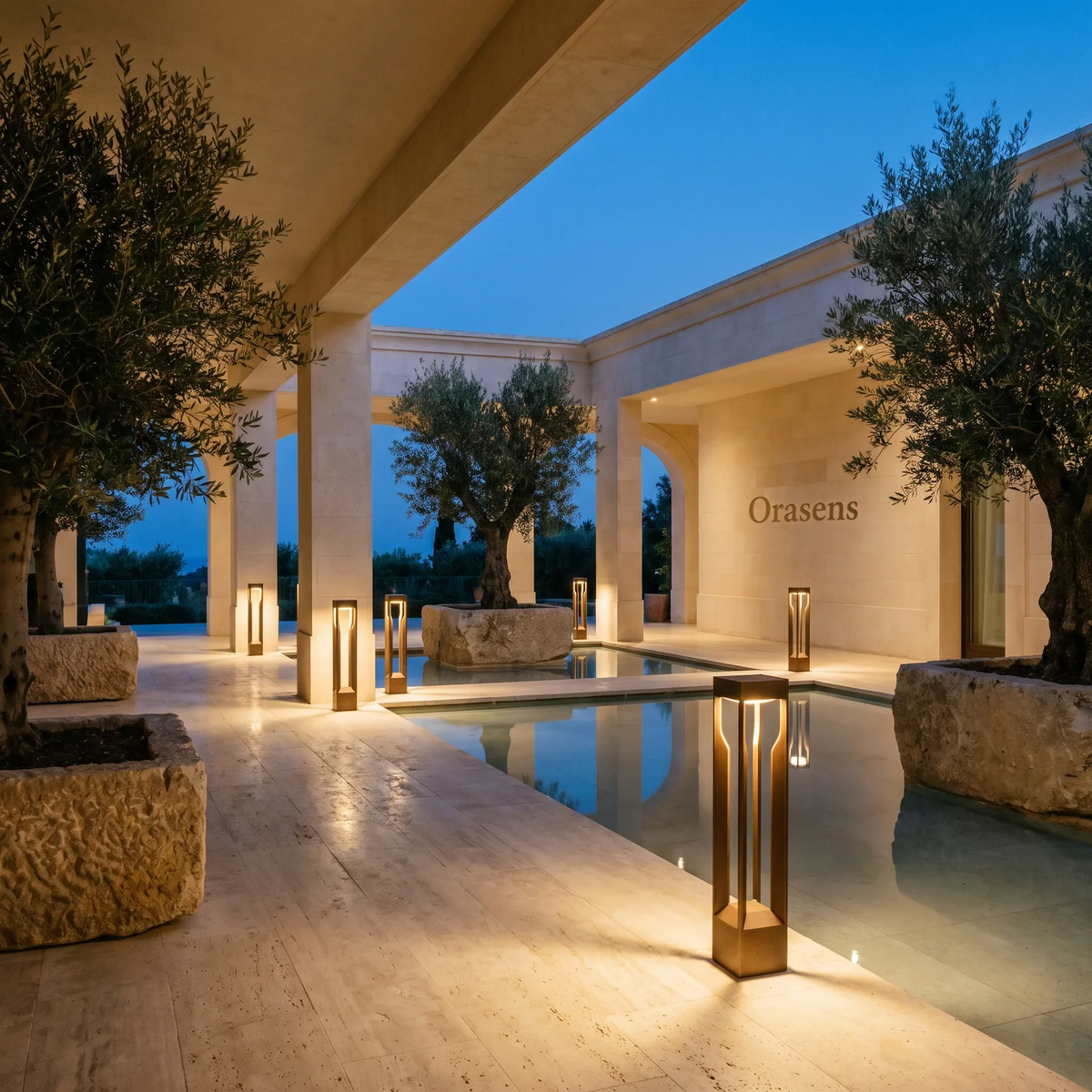 Orasens brass bollard lights along a travertine terrace and reflecting pool at dusk, with the Orasens name engraved into the limestone wall