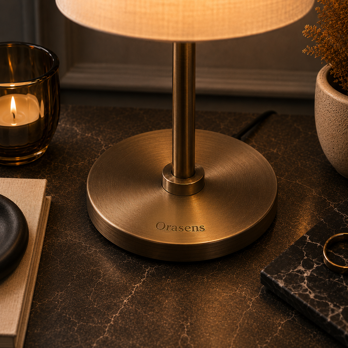 Close view of a brushed brass lamp base engraved with the Orasens name, on a dark marble table beside a lit candle
